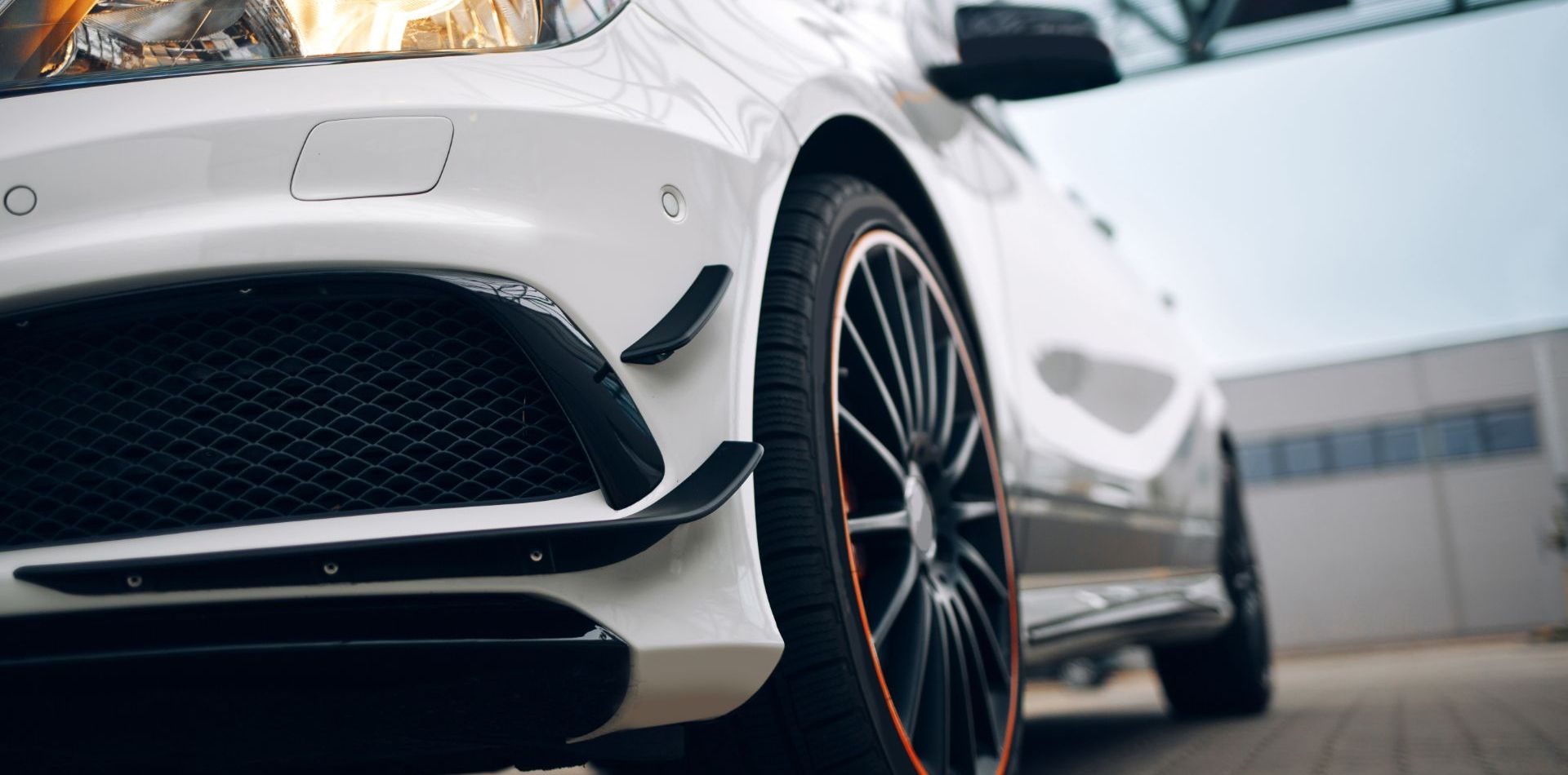 Nahaufnahme der Front eines weißen Autos mit sportlichem Design, Fokus auf Stoßfänger, Kühlergrill und Felge mit orangefarbenem Akzent